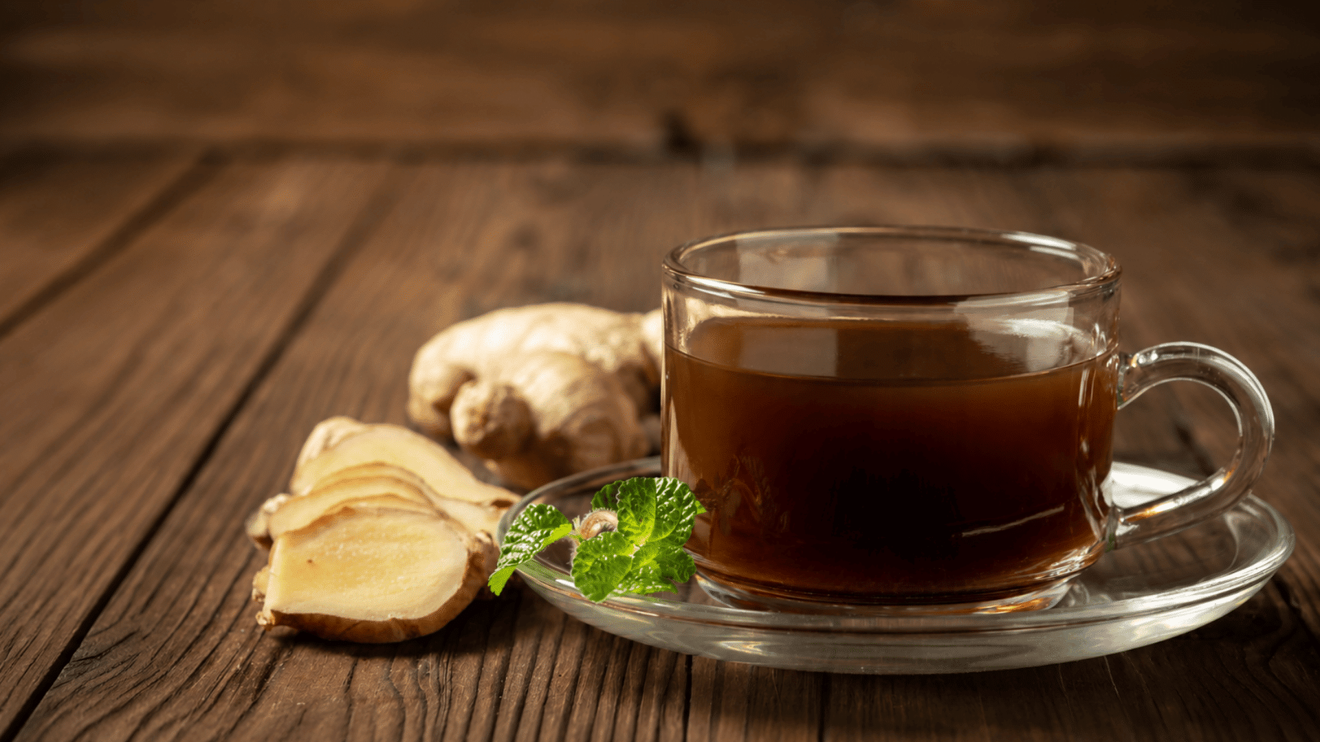a clear glass cup of ginger tea on a saucer with fresh ginger slices and mint leaves resting on a rustic wooden table
