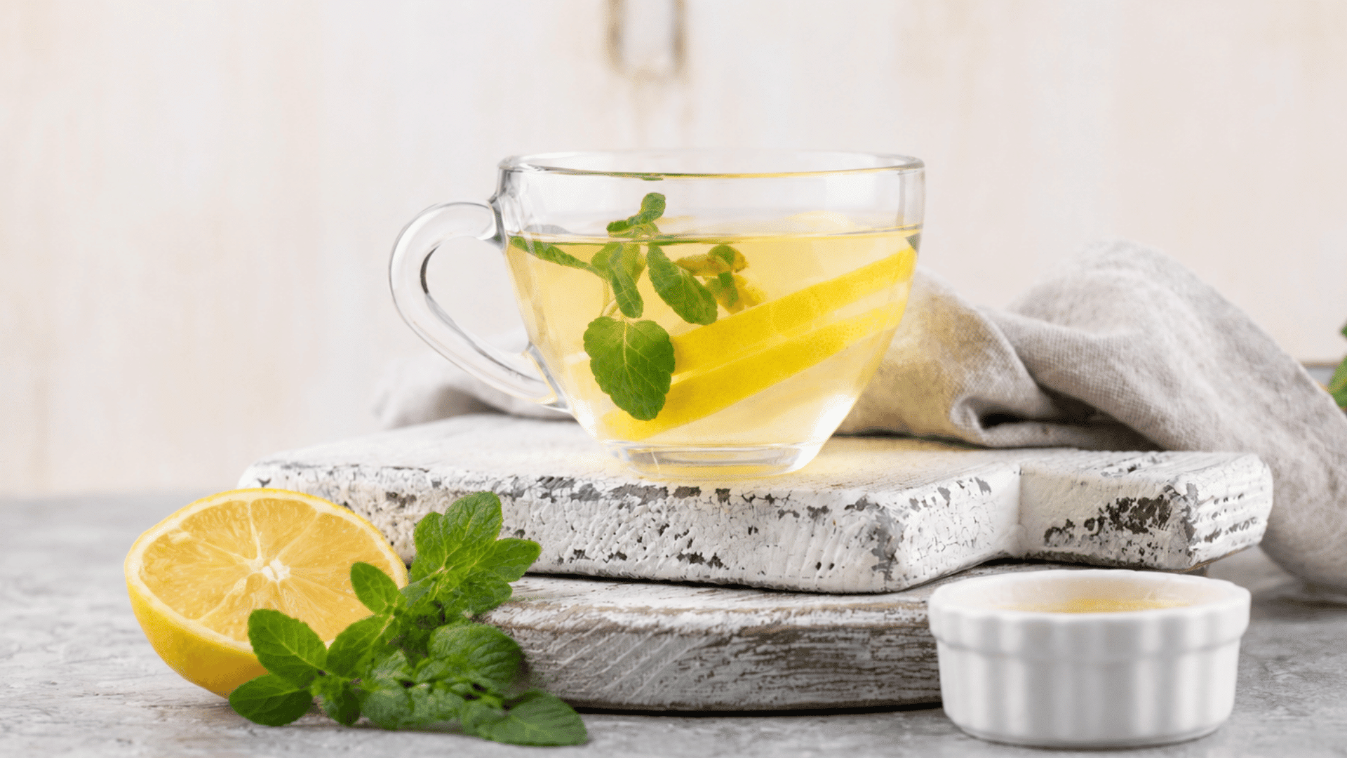 a clear glass cup of lemon tea on a rustic board with fresh lemon mint leaves and honey beside it