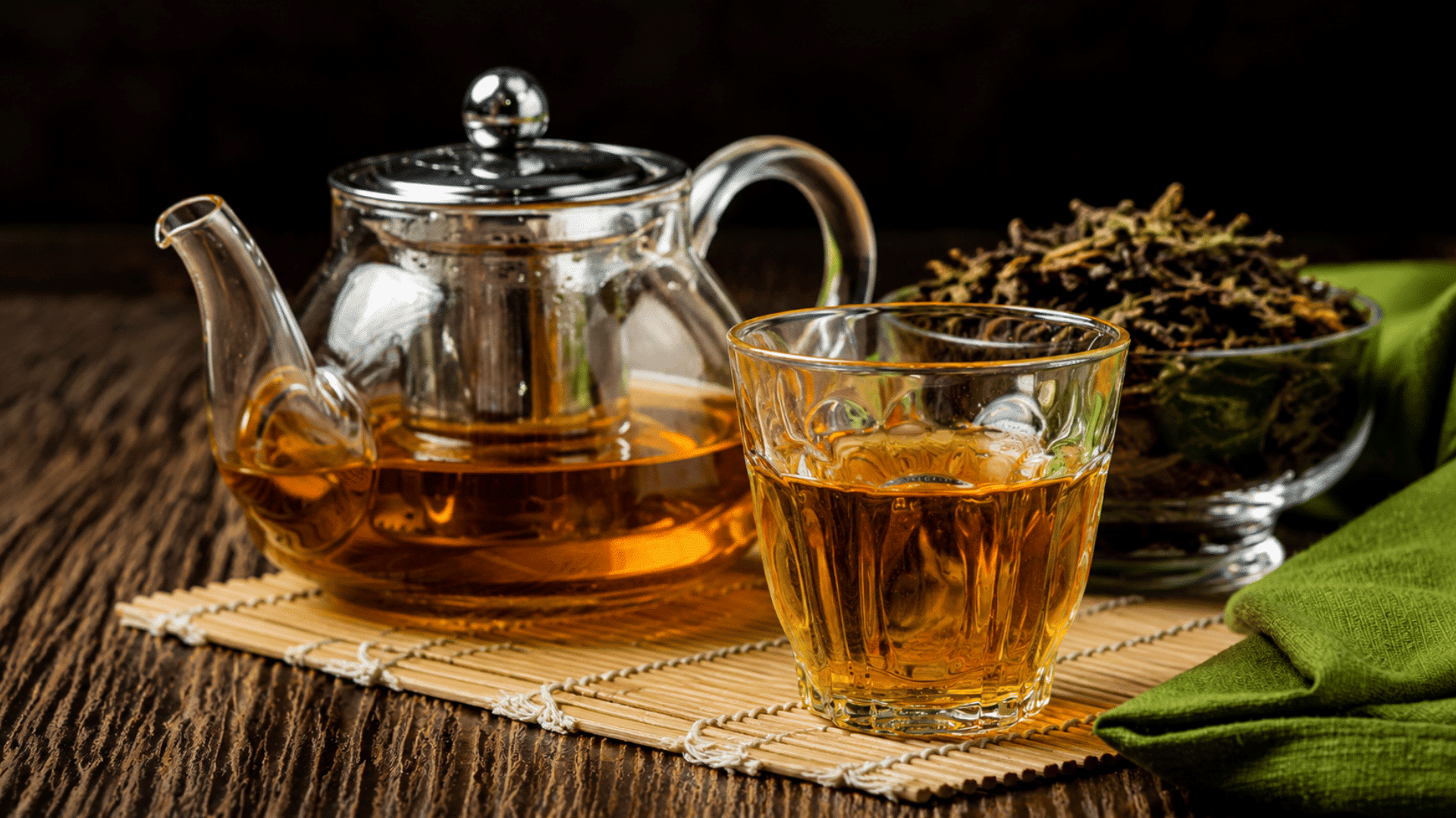a glass teapot and cup filled with golden oolong tea beside loose oolong tea leaves on a bamboo mat over a wooden table