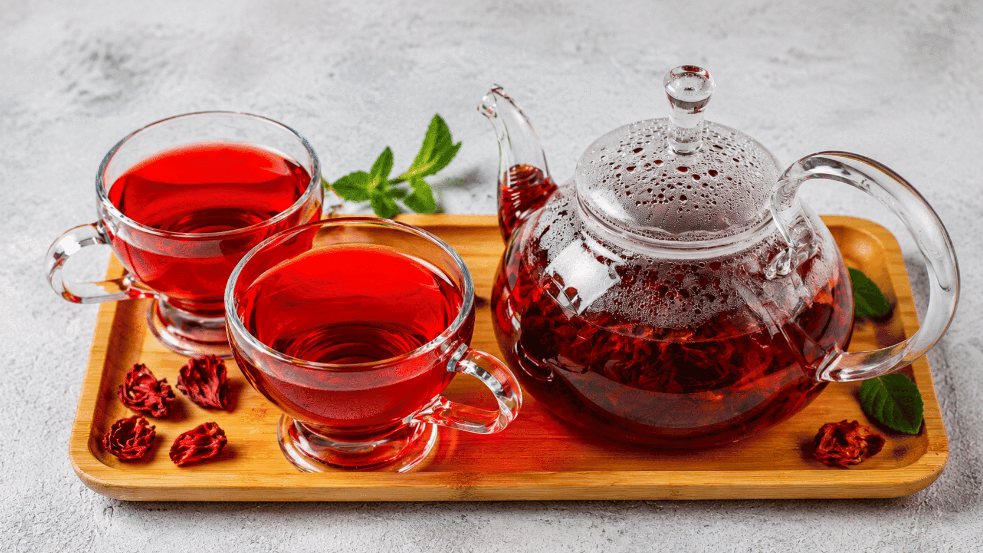 a glass teapot and two cups filled with vibrant hibiscus tea on a wooden tray with dried hibiscus flowers and fresh mint leaves