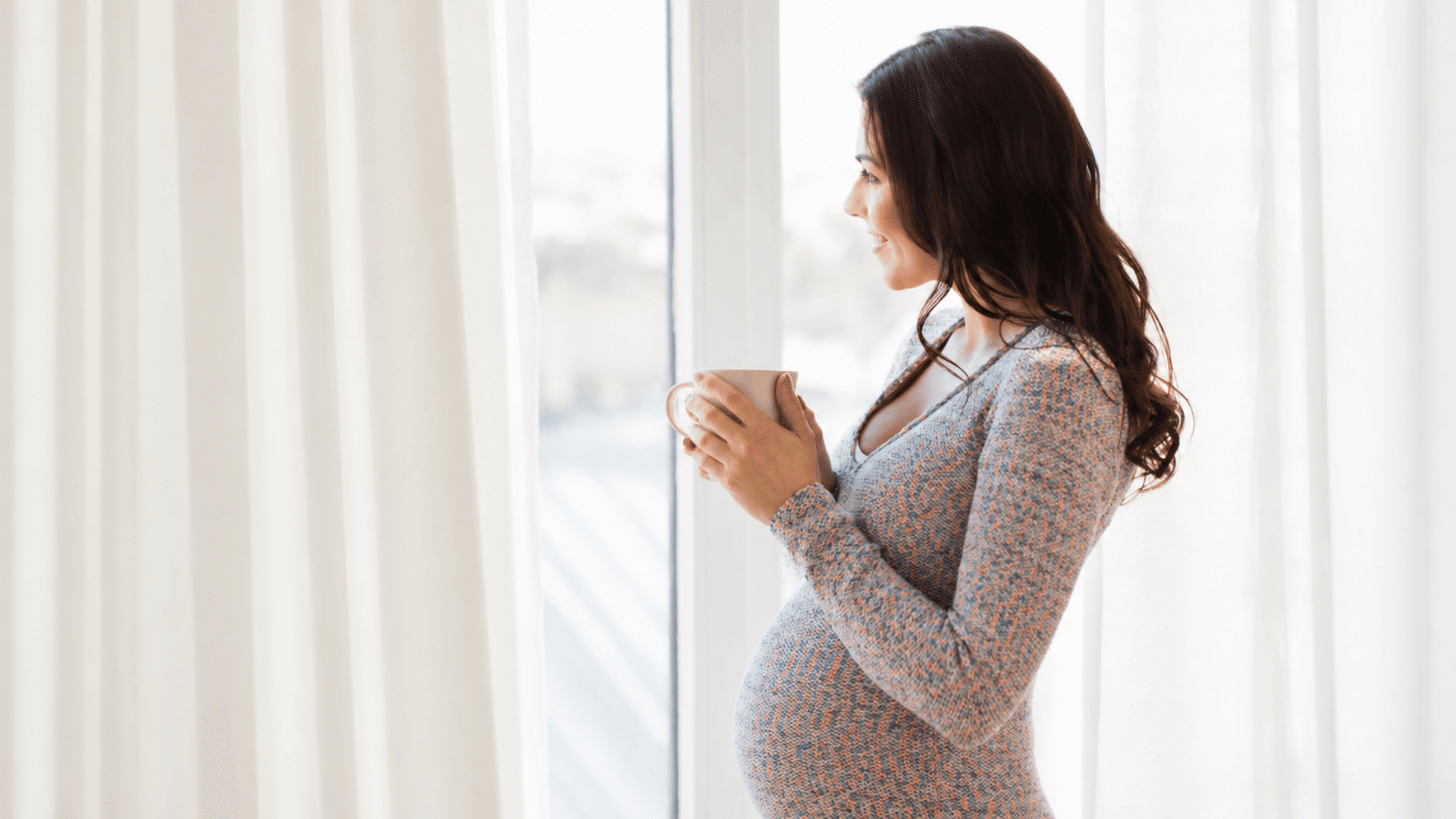 a pregnant woman holding a cup of coffee while standing by a sunlit window enjoying a quiet and relaxing moment at home