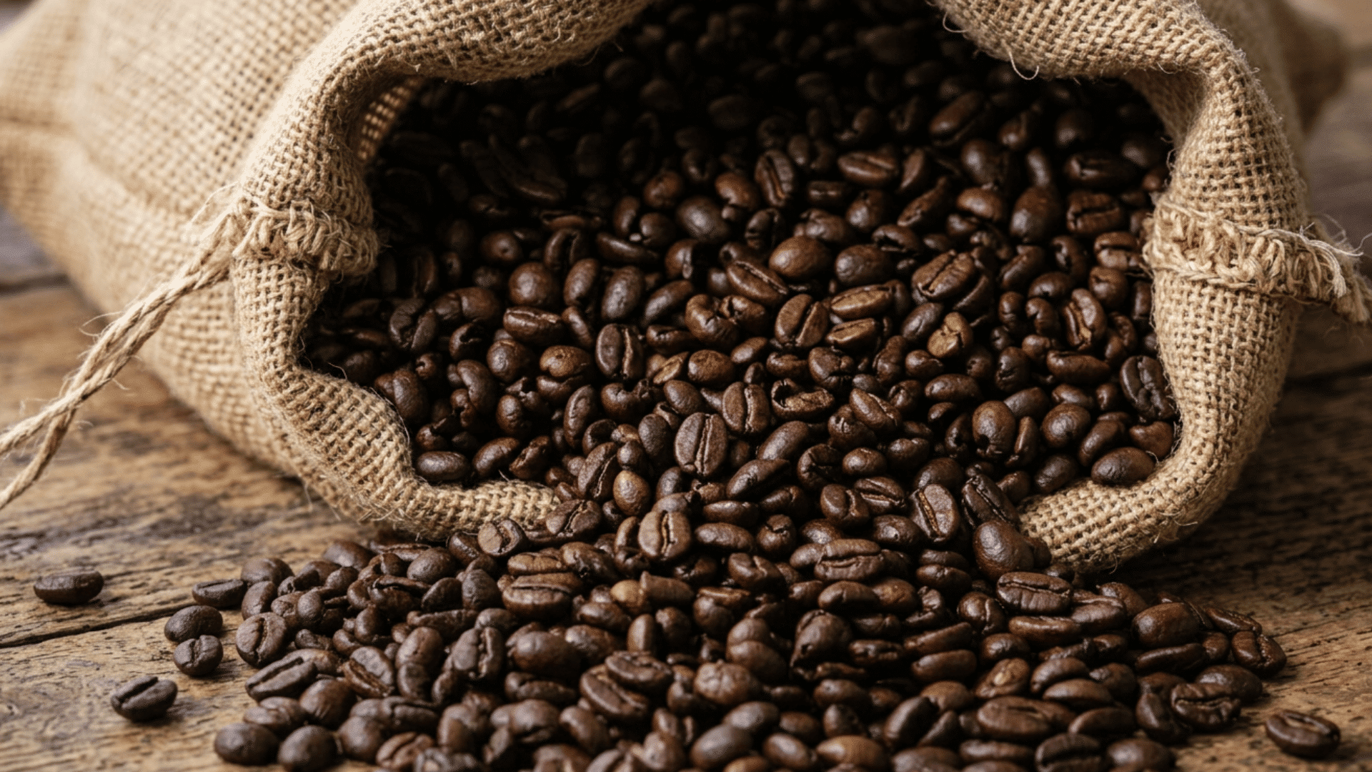 dark roasted coffee blend beans spilling from an open burlap sack onto a rustic wooden table surface