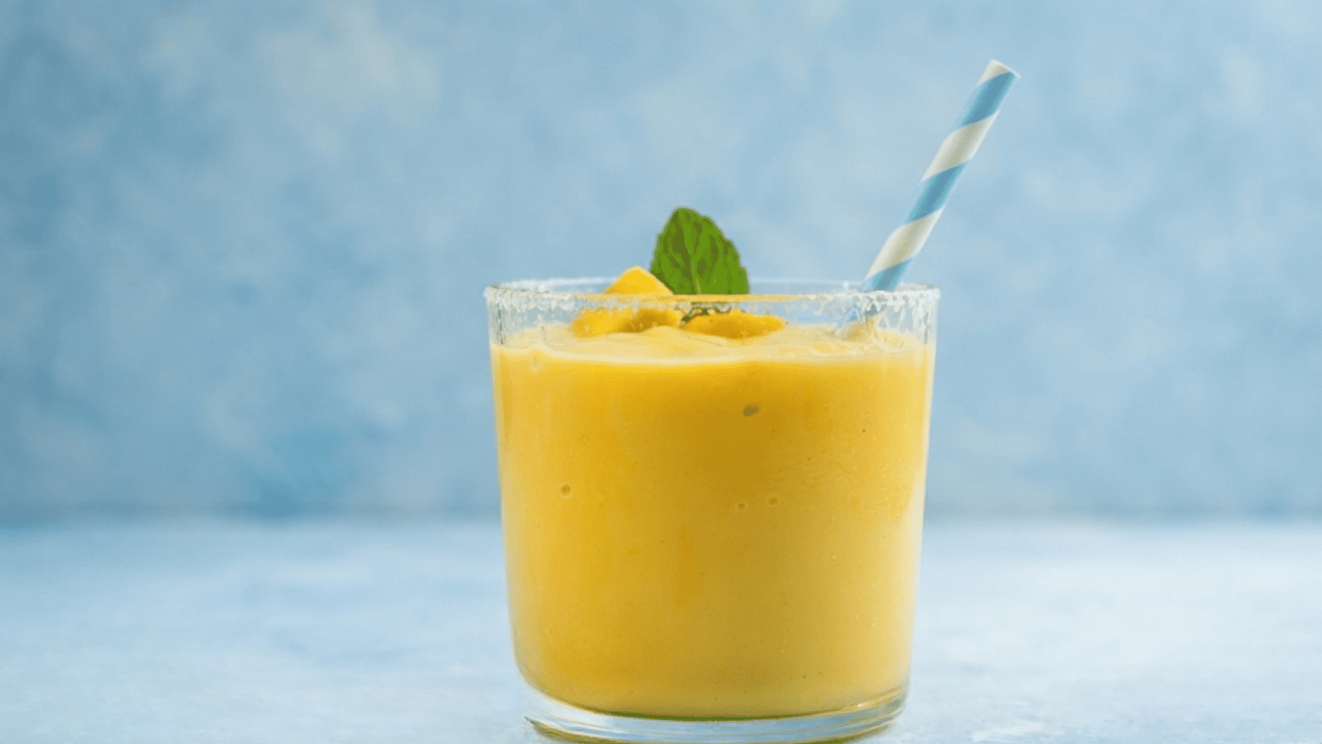 glass of creamy mango smoothie with ice topped with mint and fruit pieces and a striped paper straw on a blue surface