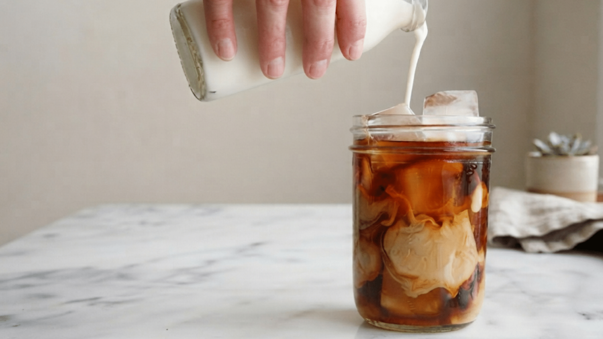 hand pouring milk into iced cold brew coffee glass jar with ice cubes on marble kitchen counter