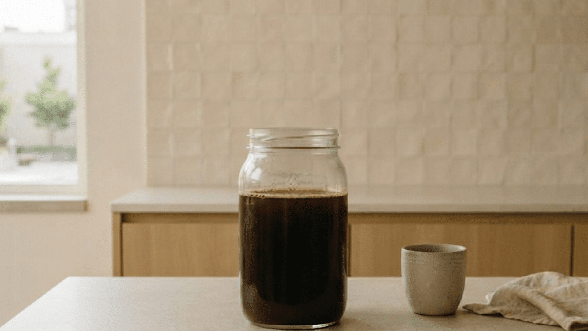 jar of cold brew coffee sitting on kitchen counter with cup and cloth beside it in a warm home setting