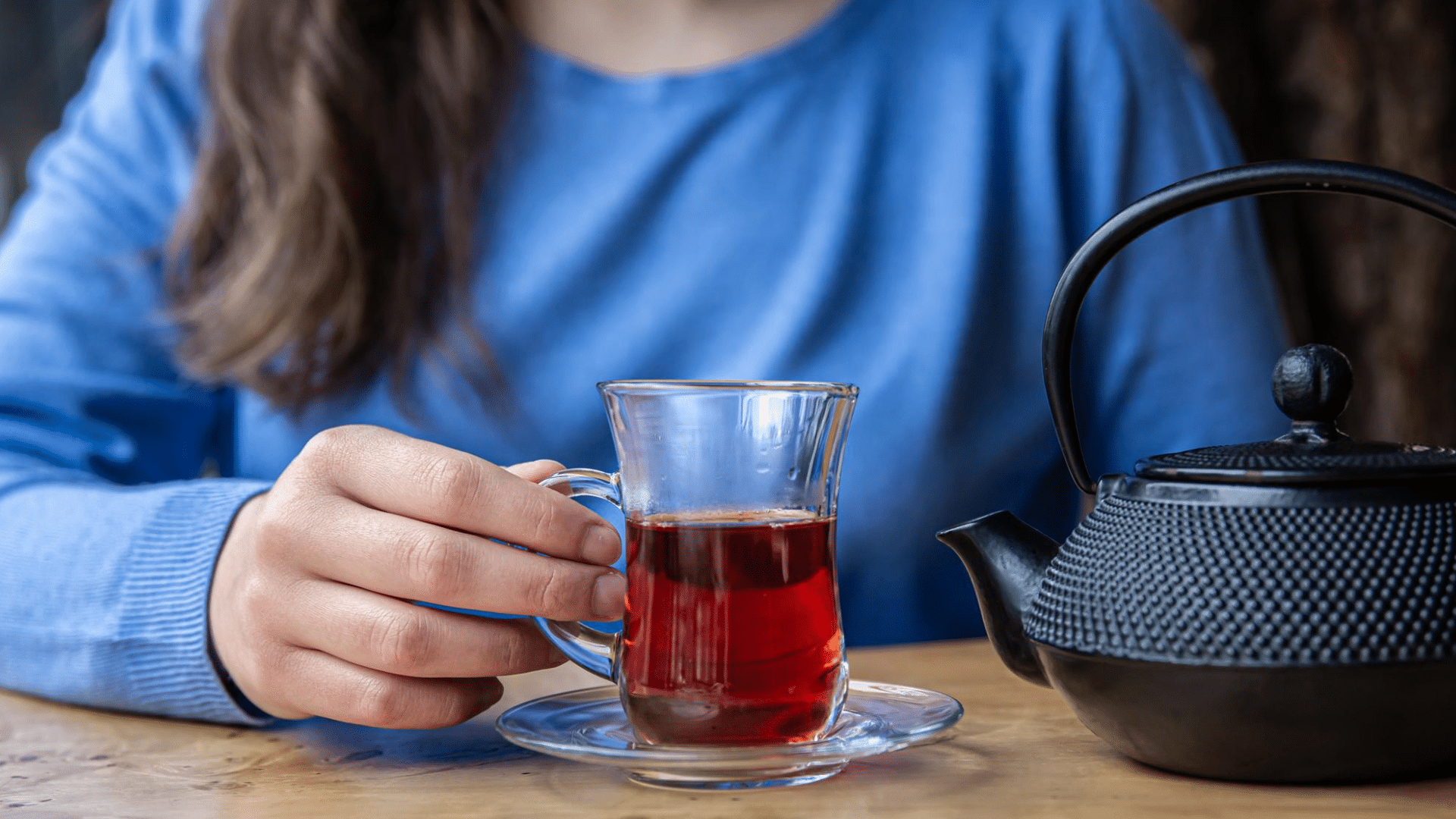 person holding a glass cup of freshly brewed black tea beside a traditional teapot on a wooden table