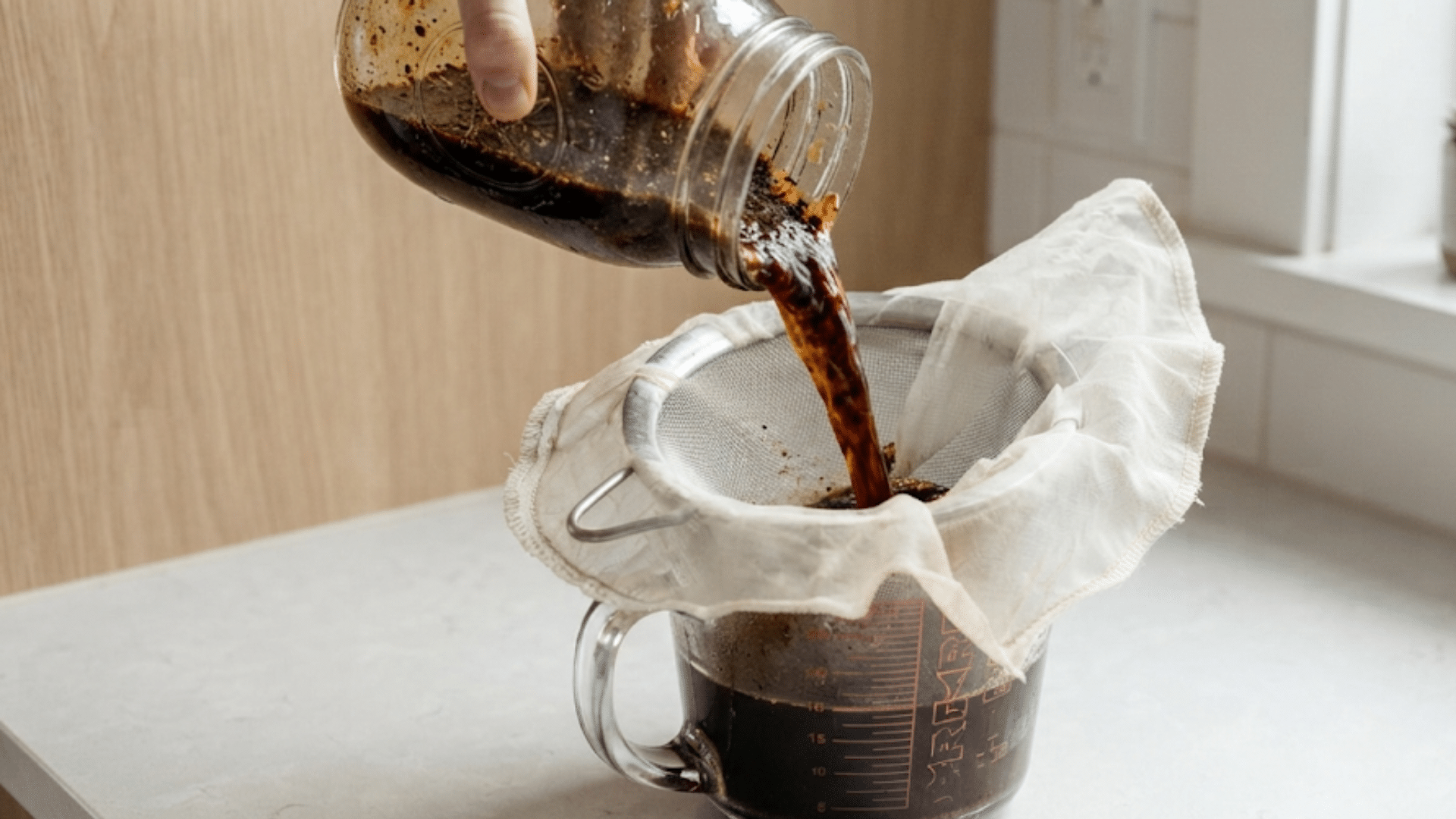 person pouring cold brew coffee through a cloth filter into glass measuring cup on kitchen counter