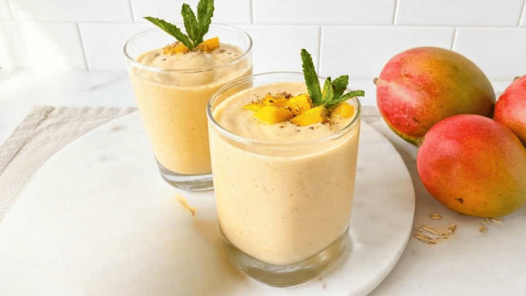 two glasses of creamy mango smoothie topped with fresh mint and mango pieces beside whole mangoes
