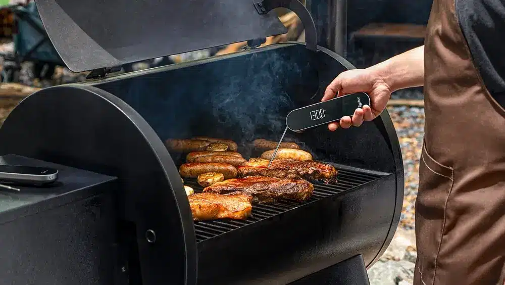 Barbecue grill with meats being cooked, person checking temperature with digital thermometer outdoors