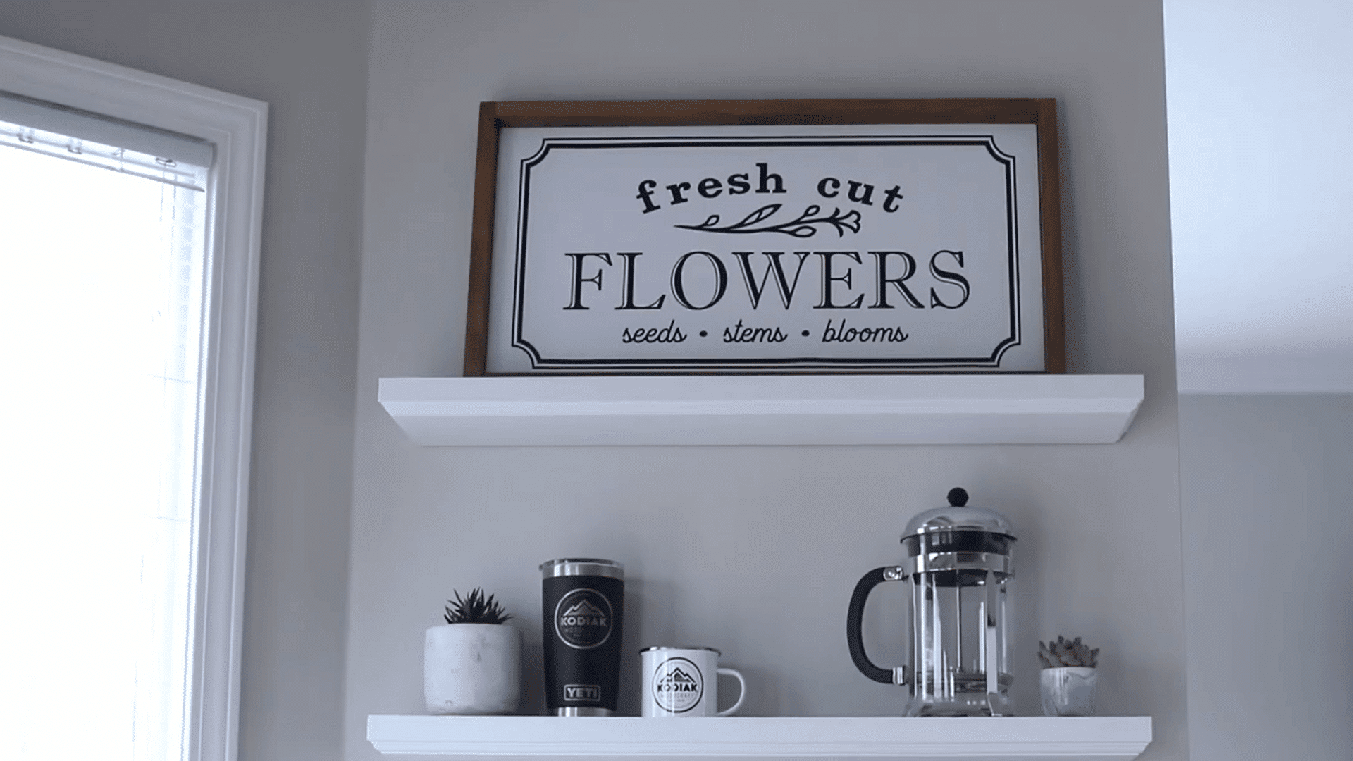 white floating shelves displaying framed flower artwork coffee accessories and small plants on a gray wall