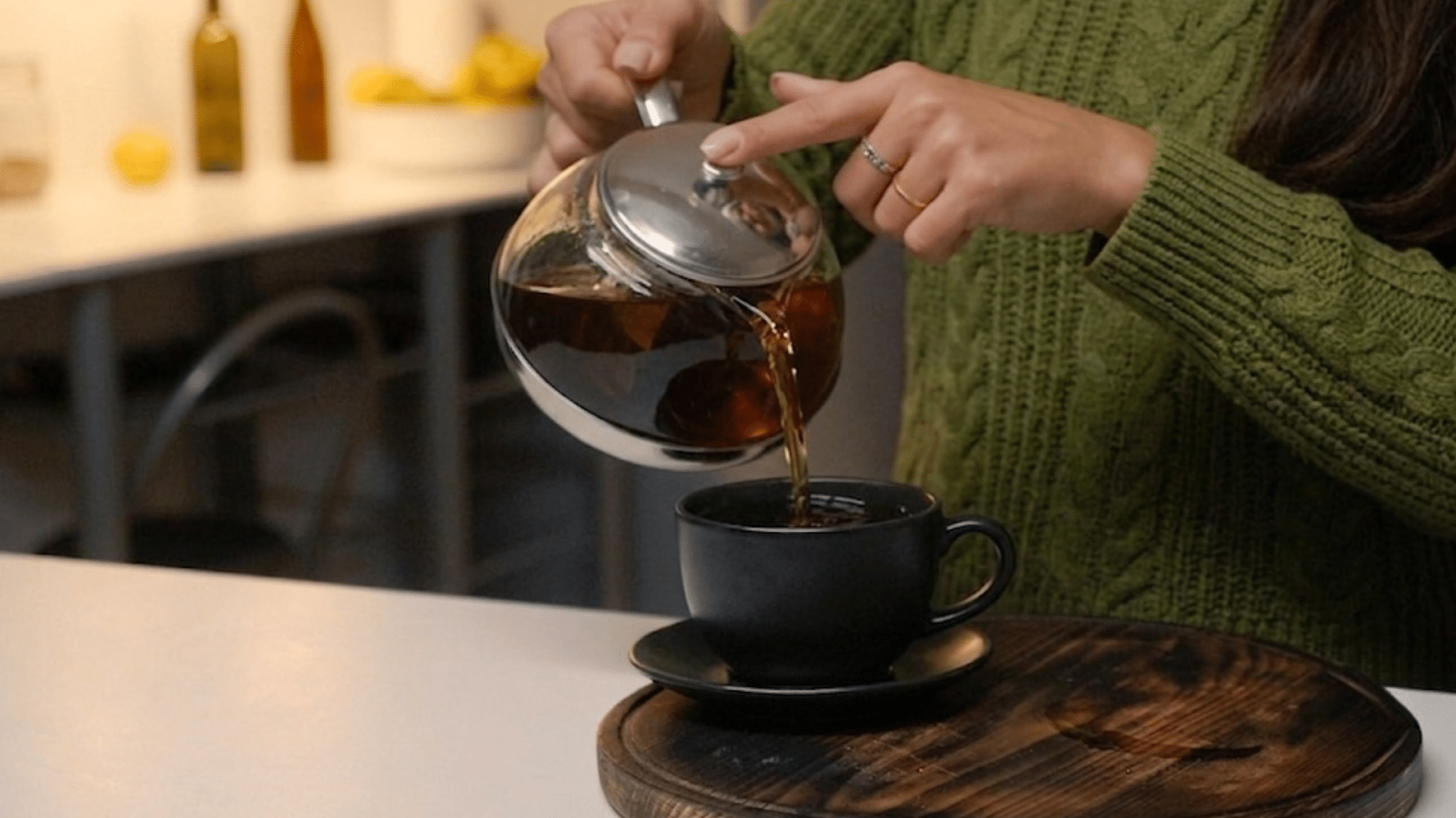 woman pouring freshly brewed black tea from a glass teapot into a black cup on a wooden tray in a cozy kitchen