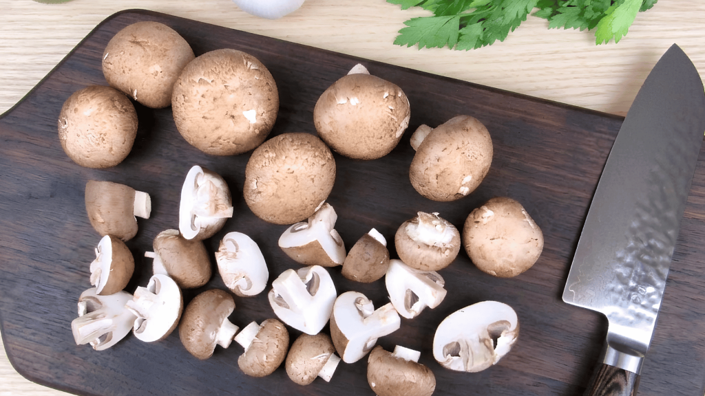 brown mushrooms whole and sliced on a wooden cutting board beside a kitchen knife and fresh parsley