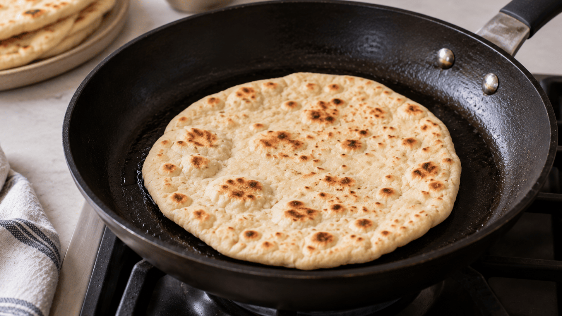flatbread cooking in a black frying pan on a stovetop with browned spots and a stack of cooked flatbreads nearby