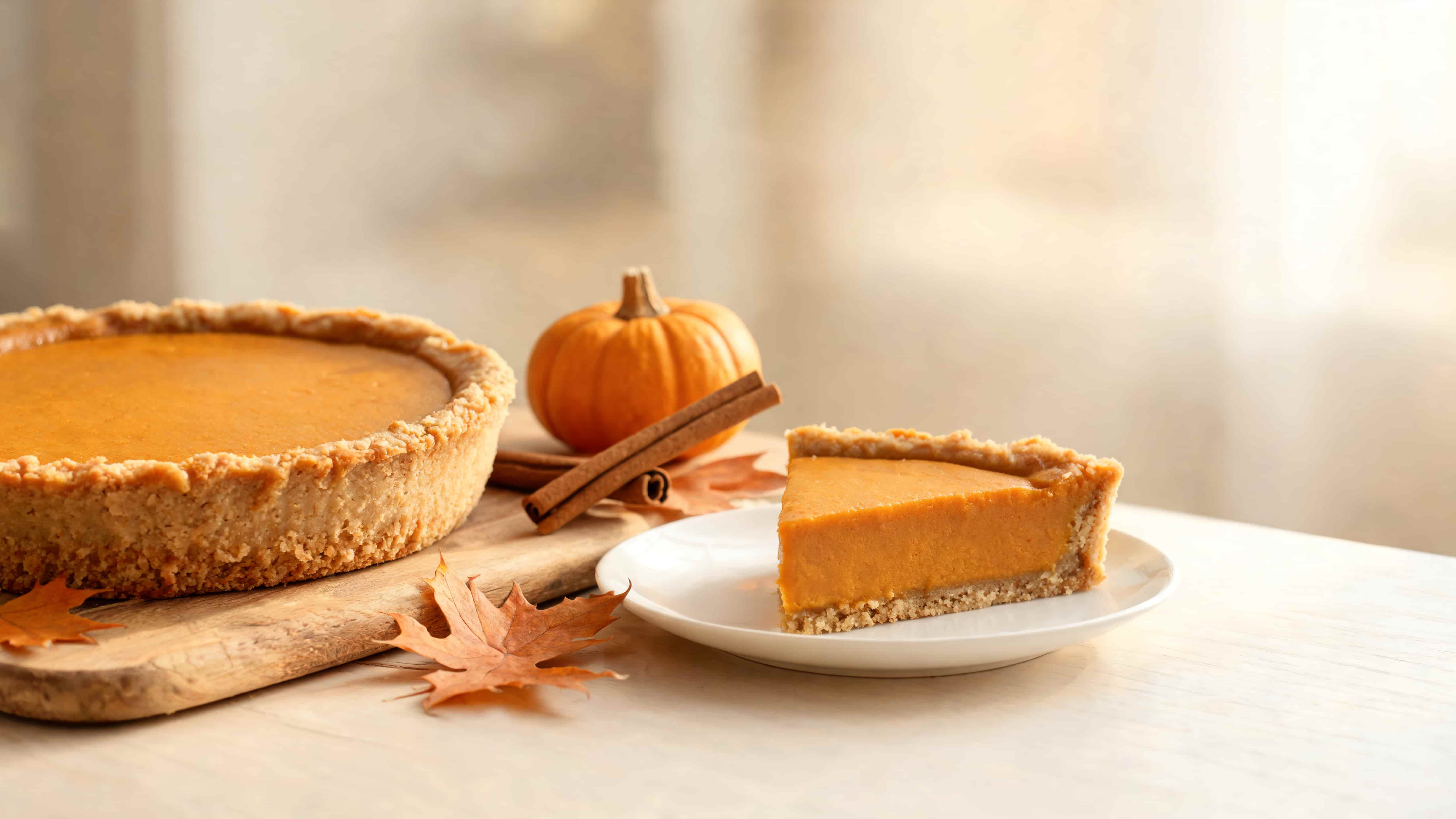 pumpkin pie with a slice served showing smooth filling, flaky crust, and autumn leaves with cinnamon sticks nearby