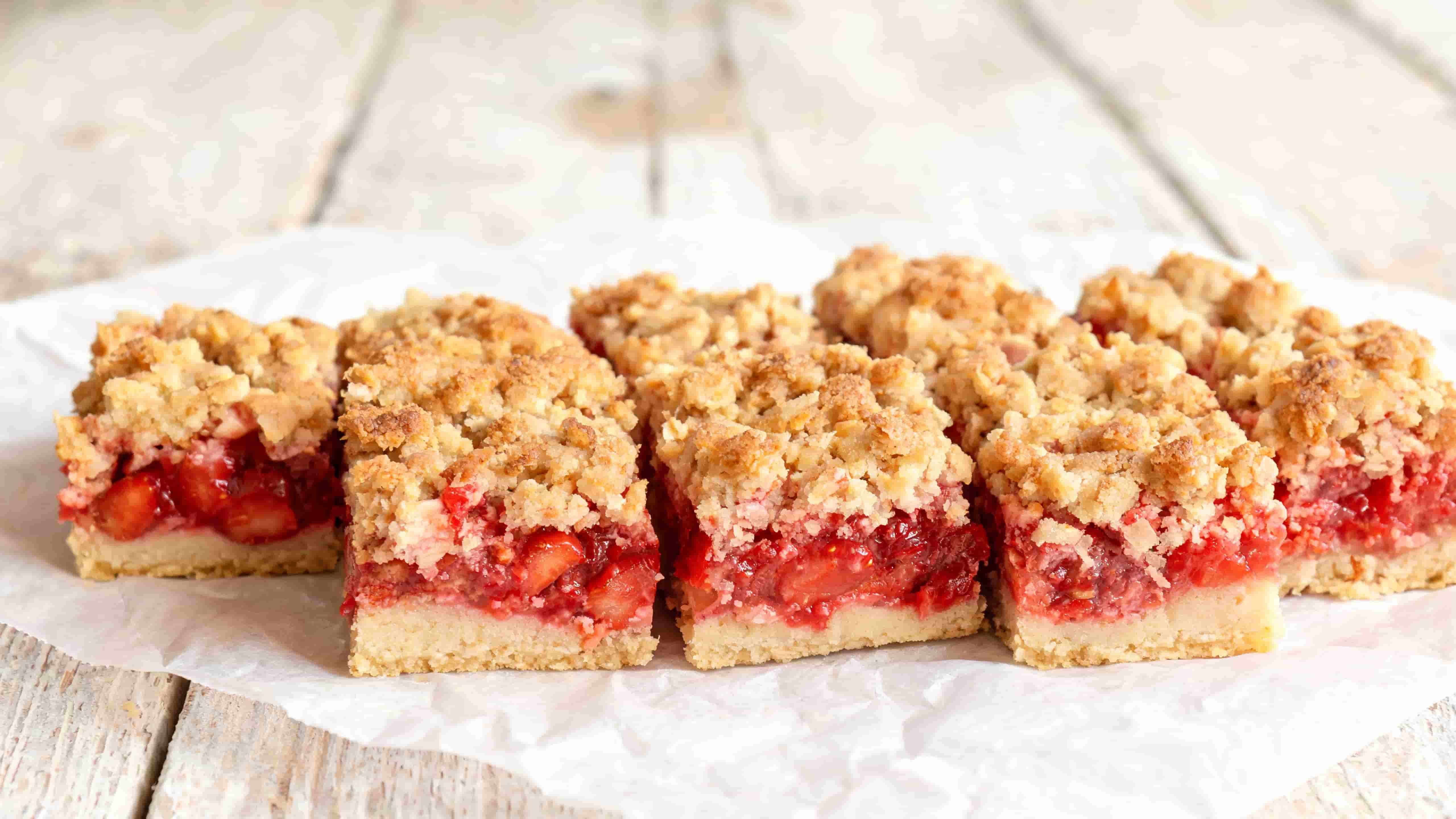 strawberry crumble bars cut into squares with visible fruit filling and golden crumb topping on parchment paper on a wooden table